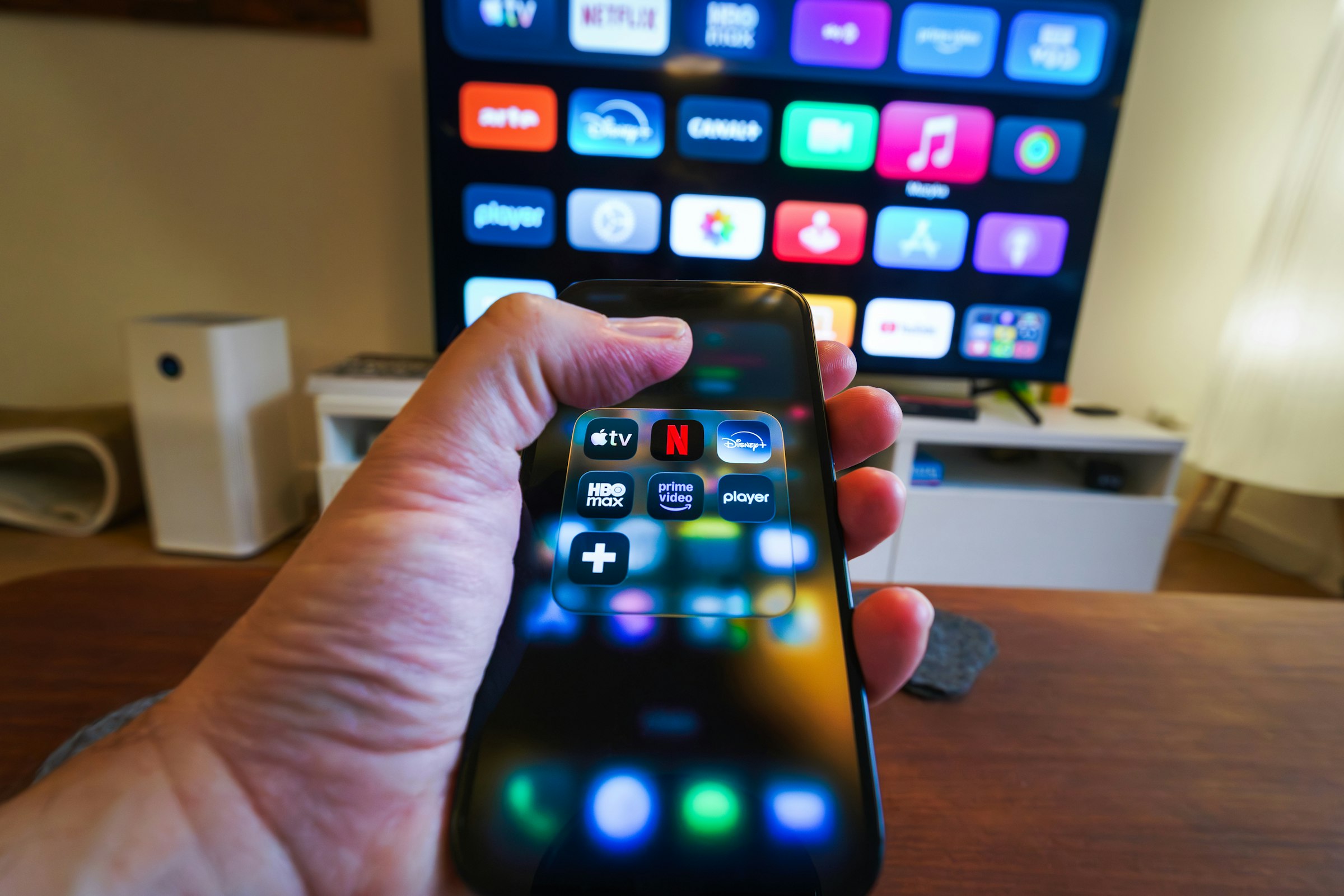 A hand holding a smartphone with streaming apps, with a TV screen blurred in the background